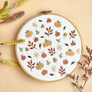 Puede incluir: Arte de bordado en aro con un fondo blanco y una variedad de elementos otoñales, incluyendo calabazas, hojas, setas y flores en tonos naranja, amarillo y marrón.