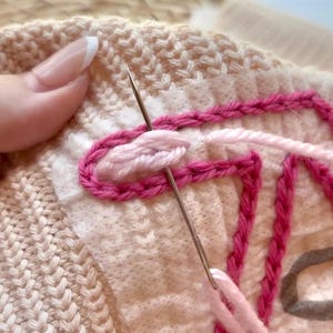 May include: Close-up of a hand embroidering a pink and white design on a knitted fabric. The needle is threaded with pink yarn and is being used to create a loop stitch.