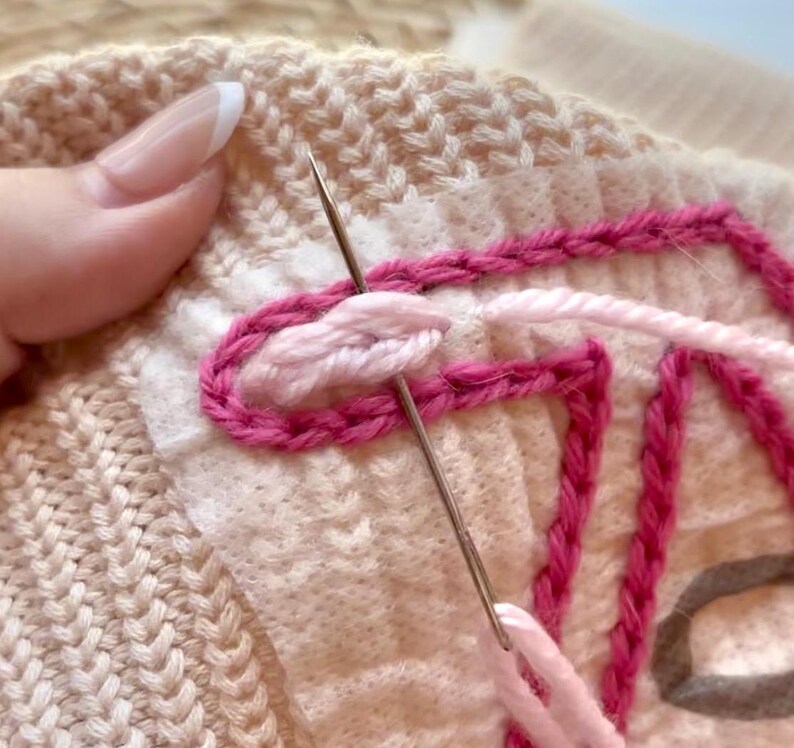 May include: Close-up of a hand embroidering a pink and white design on a knitted fabric. The needle is threaded with pink yarn and is being used to create a loop stitch.