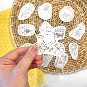 May include: A collection of white die-cut stickers featuring black line art illustrations of a bicycle with a flower basket, a bee on a honeycomb, a watering can, a sunflower, a jar labeled jam, a butterfly, and various floral motifs.