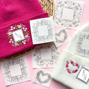 May include: A fuchsia beanie with an embroidered floral design and initials, alongside several square patches with floral and heart-shaped embroidery patterns. The patches are in various stages of completion, suggesting a DIY or craft project.