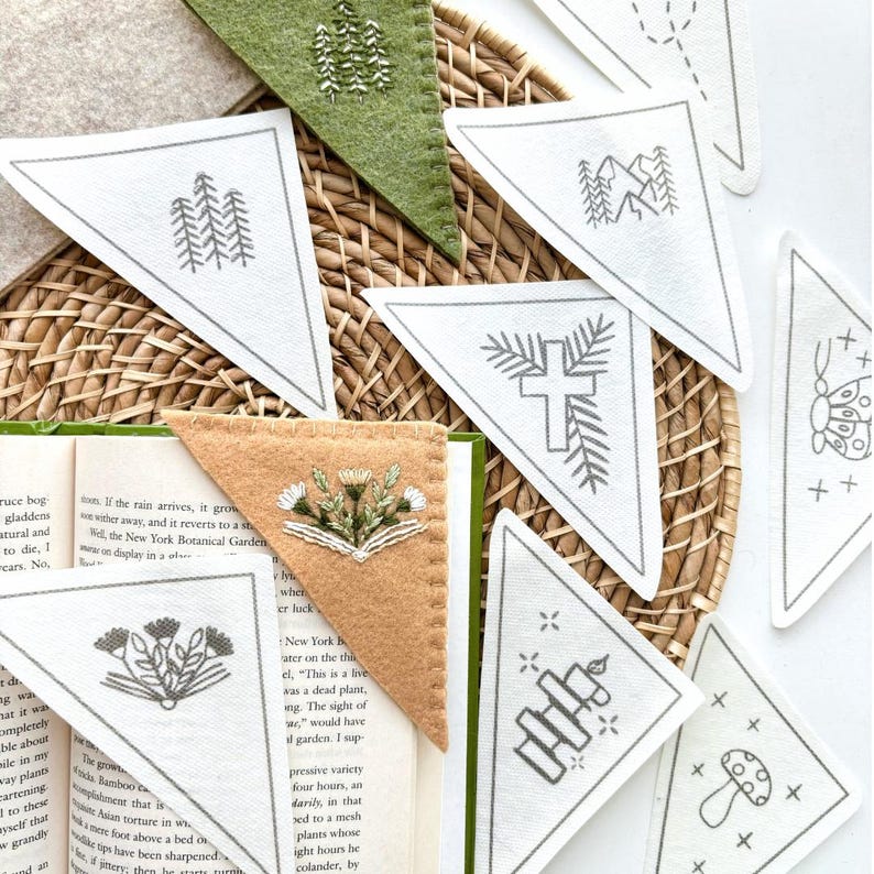 May include: A set of ten white felt triangle bookmarks with various embroidered designs, including a cross, a mushroom, a candle, a mountain range, and a butterfly. The bookmarks are arranged on a woven wicker surface.
