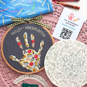May include: A dark grey embroidery hoop featuring a handprint appliqué with a school-themed pattern, embroidered with the text "PAISLEY KINDERGARTEN". Includes a white card with a QR code, a blue fabric piece with crayon prints, and a circular alphabet mat.