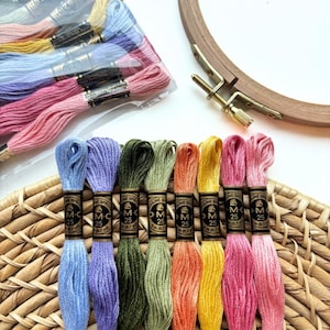 May include: A collection of colorful embroidery floss in various shades, including blue, purple, green, orange, yellow, and pink. A wooden embroidery hoop and a bundle of floss are also visible. The floss is arranged on a woven basket.