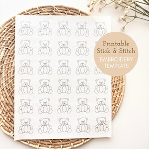 May include: A sheet of white fabric with a repeating pattern of outlined teddy bears. A tan circle with the text "Printable Stick & Stitch EMBROIDERY TEMPLATE" is on the right side. The fabric is on a woven basket.