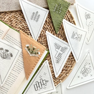 May include: A collection of triangular felt bookmarks with embroidered nature-themed designs, including trees, mountains, and a bee. The bookmarks are in various colors, including white, green, and tan, and are arranged on a woven basket.