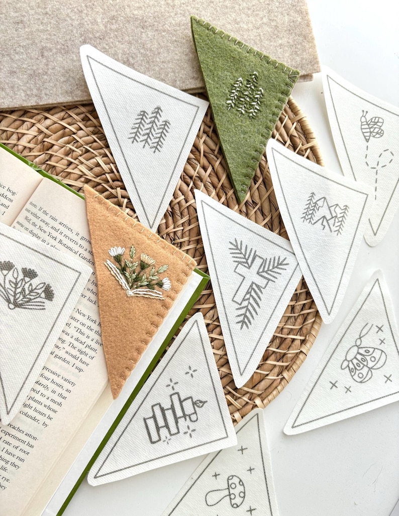 May include: A collection of triangular felt bookmarks with embroidered nature-themed designs, including trees, mountains, and a bee. The bookmarks are in various colors, including white, green, and tan, and are arranged on a woven basket.