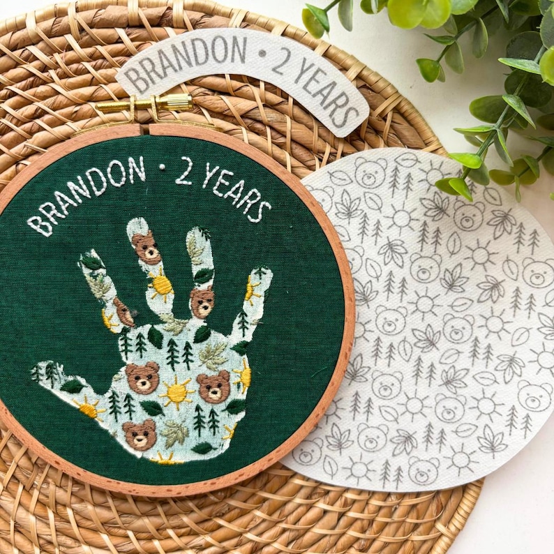 May include: An embroidered hoop art piece with a handprint design. The handprint is filled with bear, tree, and sun motifs. The text "BRANDON - 2 YEARS" is embroidered on the green fabric. A separate circular fabric piece features a repeating pattern of bears, trees, and suns.