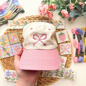 May include: A cream-colored baseball cap with a pink brim and a pink embroidered bow design. The cap is surrounded by cross-stitch samplers with floral and fruit patterns, along with spools of colorful embroidery floss. The overall theme is crafting and handmade items.