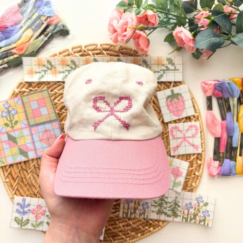 May include: A cream-colored baseball cap with a pink brim and a pink embroidered bow design. The cap is surrounded by cross-stitch samplers with floral and fruit patterns, along with spools of colorful embroidery floss. The overall theme is crafting and handmade items.