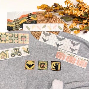 May include: A grey sweatshirt featuring three small cross-stitch squares of a bat, a pumpkin, and a black cat. Several paper cross-stitch patterns are arranged on the fabric, including designs of leaves, bats, pumpkins, a tractor, and a barn.