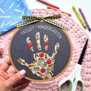 May include: A circular wooden embroidery hoop containing dark gray fabric with a handprint shape filled with school-themed patterns including apples, pencils, and rainbows. The text "PAISLEY &bull; KINDERGARTEN" is stitched above, topped with a green gingham bow.