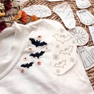 K&ouml;nnte beinhalten: Ein wei&szlig;es T-Shirt mit schwarzen gestickten Flederm&auml;usen und kleinen rosa Blumen am Halsausschnitt. Mehrere wei&szlig;e Stoffaufn&auml;her mit schwarzen Linienzeichnungen von Flederm&auml;usen, Blumen, einem Geist, einer Katze und einem K&uuml;rbis liegen darauf.