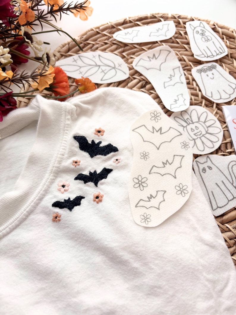 K&ouml;nnte beinhalten: Ein wei&szlig;es T-Shirt mit schwarzen gestickten Flederm&auml;usen und kleinen rosa Blumen am Halsausschnitt. Mehrere wei&szlig;e Stoffaufn&auml;her mit schwarzen Linienzeichnungen von Flederm&auml;usen, Blumen, einem Geist, einer Katze und einem K&uuml;rbis liegen darauf.