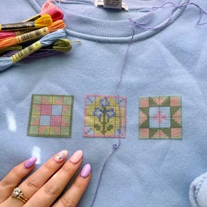 May include: Light blue sweatshirt with three embroidered quilt block designs in pink, green, and yellow. Embroidery floss in various colors and a hand with painted nails are also visible.