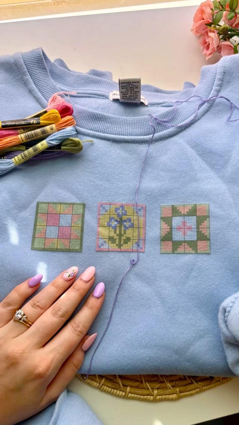 May include: Light blue sweatshirt with three embroidered quilt block designs in pink, green, and yellow. Embroidery floss in various colors and a hand with painted nails are also visible.