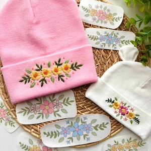 May include: A pink knit beanie featuring an embroidered yellow floral design and a white knit beanie with a multi-colored floral embroidery, accompanied by several white fabric patches displaying various colorful floral patterns.