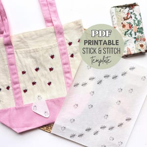 May include: A cream-colored tote bag with pink accents and handles, adorned with red ladybug embroidery. A printed template with ant and ladybug designs is next to the bag. The image also includes a fabric roll and a sticker.