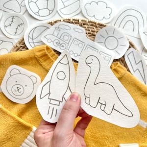 May include: A set of iron-on patches featuring various designs, including a rocket, dinosaur, bear, train, sun, rainbow, and football. The patches are white with black outlines and are ready to be ironed onto fabric.