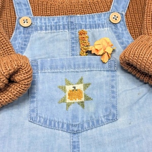Puede incluir: Peto vaquero azul claro con un bolsillo delantero que presenta un diseño de estrella y calabaza en punto de cruz. El peto tiene dos botones claros en la parte superior y se lleva sobre un suéter de punto grueso marrón.