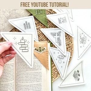 May include: A set of nine white felt triangle bookmarks with various embroidered designs, including a cross, a mushroom, a butterfly, a candle, a tree, and a flower. The bookmarks are arranged on a woven wicker surface with a book and a green felt bookmark in the background. The text "FREE YOUTUBE TUTORIAL!" is at the top of the image.