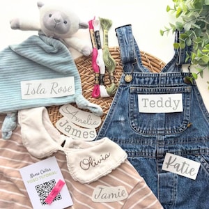 May include: A light blue ribbed hat, a denim overall, a beige striped shirt with a white collar embroidered with the name Olivia, a plush lamb, three embroidery floss skeins, and several white labels printed with the names Isla Rose, Teddy, Amelia, James, Katie, and Jamie.
