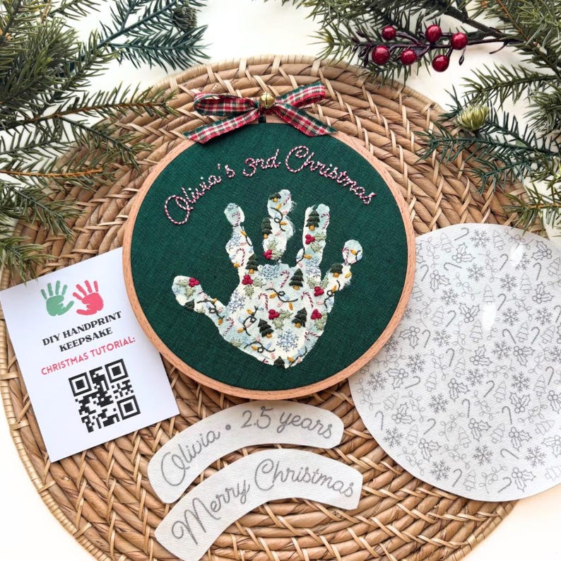 Puede incluir: Un aro de bordado de tela verde con un dise&ntilde;o de huella de mano, luces navide&ntilde;as festivas y el texto "Olivia's 3rd Christmas." Incluye un tutorial y dos etiquetas de tela que dicen "Olivia 25 a&ntilde;os" y "Feliz Navidad."