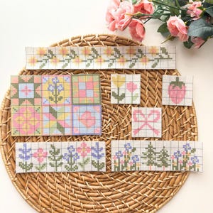 May include: A collection of cross-stitch patterns featuring floral and geometric designs in pastel colors. The patterns include flowers, a strawberry, and a bow, arranged on a woven basket. Pink roses are in the background.