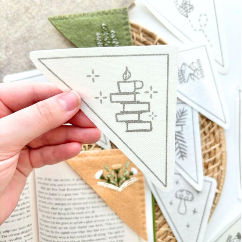 May include: A set of white felt triangle bookmarks with different hand-drawn illustrations. The bookmark in the foreground features a stack of books with a candle on top. The other bookmarks feature a mushroom, a pine tree, a book with flowers, and a bee.