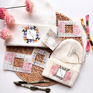 May include: Two white knit beanies featuring cross-stitch floral patterns, accompanied by paper cross-stitch templates, embroidery thread in pink and olive green, and a needle. One beanie displays a floral wreath with the letters SWS.