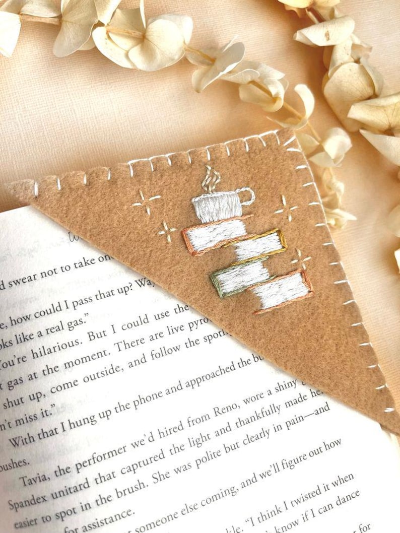 May include: A tan felt bookmark with white stitching and embroidered details. The design features a stack of books topped with a steaming coffee cup. The bookmark is placed on the corner of a book with printed text.