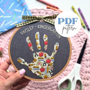 May include: A hand-shaped embroidery hoop with the text "Paisley Kindergarten" and a "PDF pattern" label. The handprint design features colorful school-themed elements like pencils, apples, and rainbows. A green and brown checkered ribbon is tied at the top.