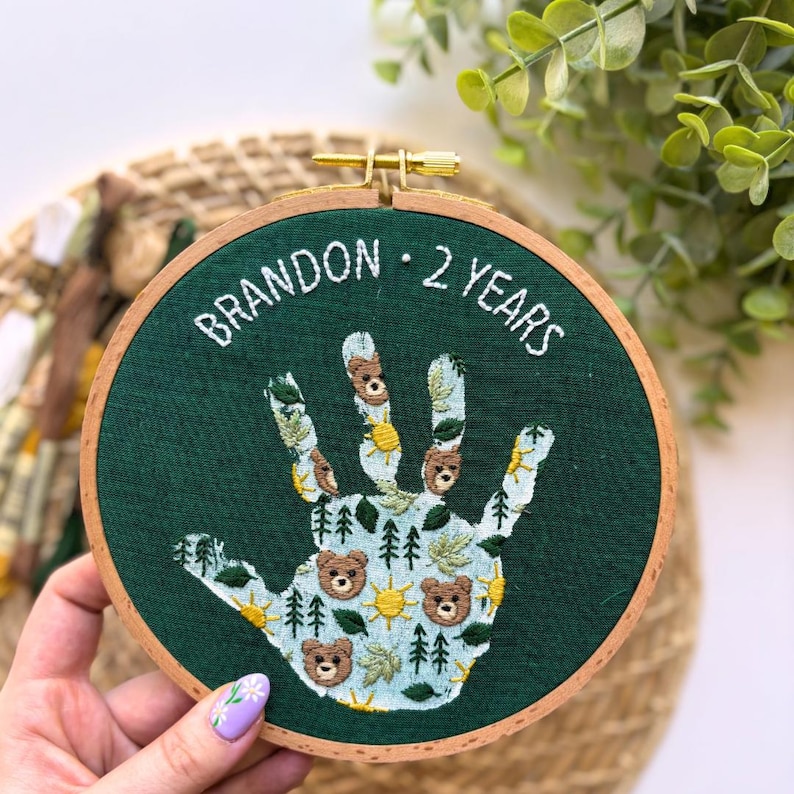 May include: An embroidered handprint design on a dark green fabric, framed in a wooden hoop. The design includes bears, trees, and suns, with the text "BRANDON &bull; 2 YEARS" stitched in white thread. The hoop is held by a person with a painted fingernail.