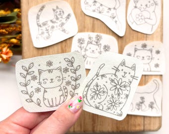 Può includere: Una collezione di toppe a tema gatto con disegni floreali. Le toppe presentano varie pose di gatti e sono decorate con fiori. Le toppe sono grigie e bianche e sono tenute in mano con un design floreale per unghie.