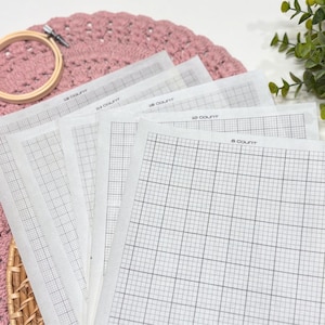 May include: A set of white cross stitch fabric sheets with black grid lines, each labelled with its count including 6, 10, 14, 16, and 18 count. The sheets are stacked and arranged with a wooden embroidery hoop and a pink crocheted mat visible.