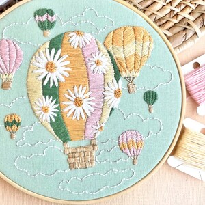 May include: A green, pink, and yellow hot air balloon with white daisy embroidery floats in a blue sky with white clouds.  The balloon is surrounded by smaller hot air balloons in various colors.