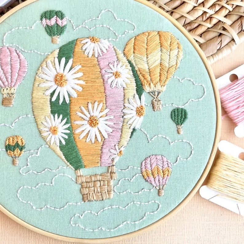 May include: A green, pink, and yellow hot air balloon with white daisy embroidery floats in a blue sky with white clouds.  The balloon is surrounded by smaller hot air balloons in various colors.