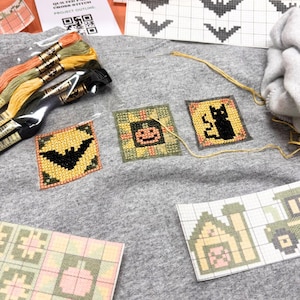May include: A gray fabric surface featuring three cross-stitch squares depicting a black bat, an orange pumpkin, and a black cat. Included are bundles of orange, green, and yellow embroidery thread, a needle, and printed cross-stitch patterns.