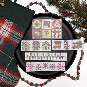 May include: A dark green plaid Christmas stocking next to a dark brown plate featuring several cross-stitch pattern charts. The charts display holiday motifs including a nutcracker, Christmas tree, houses, cars with trees, string lights, and snowflakes.
