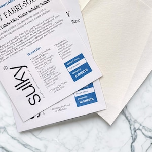May include: White Fabri-Solvy stabilizer sheets for embroidery and quilting. The image shows two packages of the water-soluble, fabric-like stabilizer, with text and product details. One package contains 6 sheets, and the other contains 12 sheets.