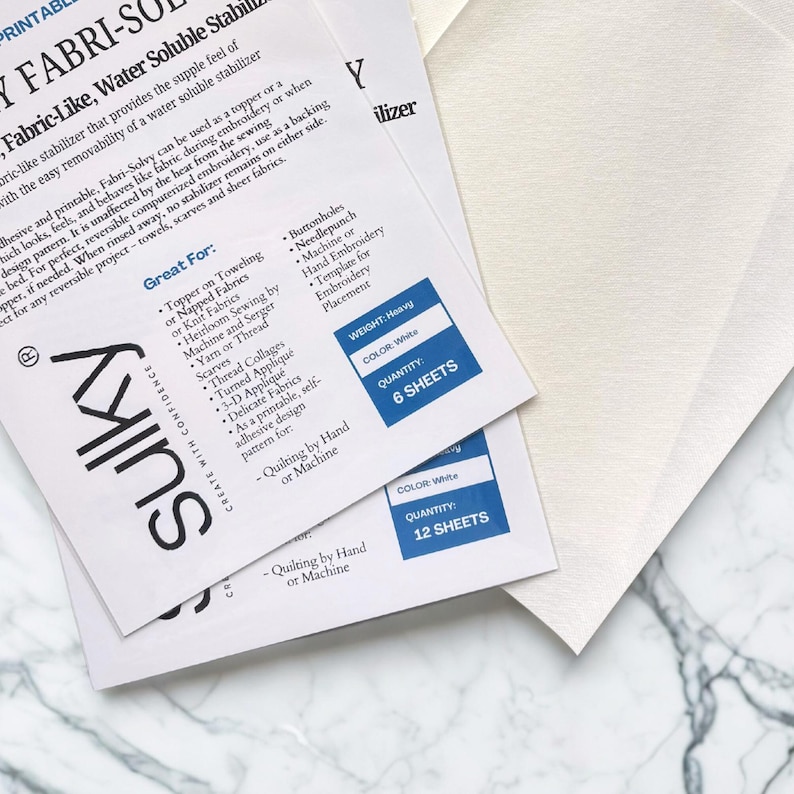 May include: White Fabri-Solvy stabilizer sheets for embroidery and quilting. The image shows two packages of the water-soluble, fabric-like stabilizer, with text and product details. One package contains 6 sheets, and the other contains 12 sheets.