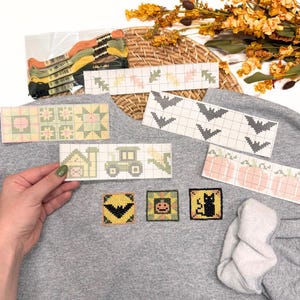 May include: A gray sweatshirt featuring three small square cross-stitch patches of a bat, a pumpkin, and a black cat. Several paper cross-stitch patterns are displayed on top, showing designs of leaves, bats, a farm, a tractor, and pumpkins.