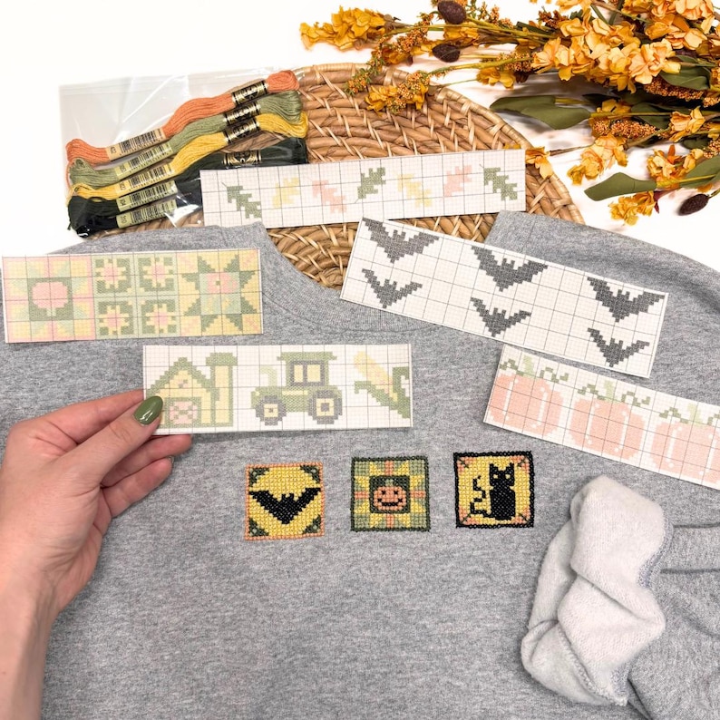 May include: A gray sweatshirt featuring three small square cross-stitch patches of a bat, a pumpkin, and a black cat. Several paper cross-stitch patterns are displayed on top, showing designs of leaves, bats, a farm, a tractor, and pumpkins.