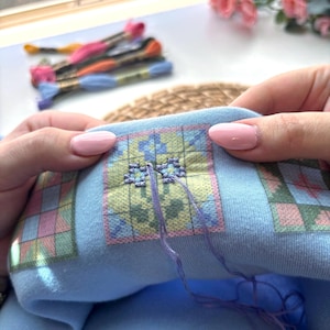 May include: Close-up of a cross-stitch project in progress. The light blue fabric features a colorful geometric pattern. The needle is threaded with purple floss, and several skeins of embroidery floss in various colors are visible in the background.