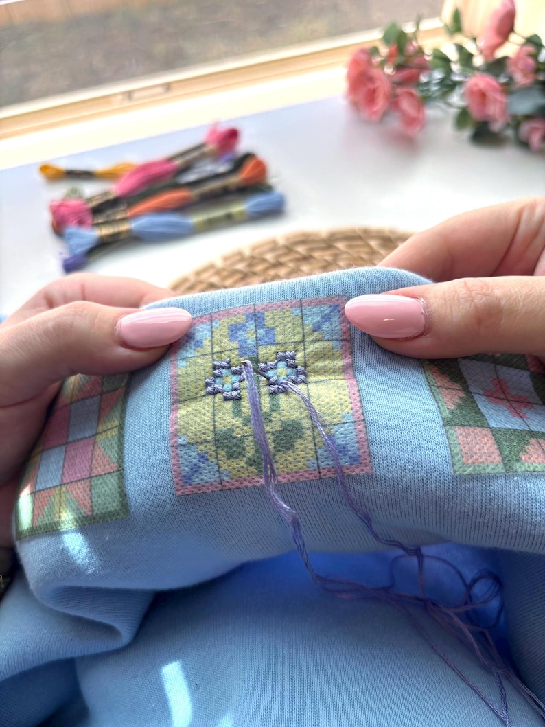 May include: Close-up of a cross-stitch project in progress. The light blue fabric features a colorful geometric pattern. The needle is threaded with purple floss, and several skeins of embroidery floss in various colors are visible in the background.
