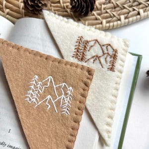 May include: Two triangular felt bookmarks. One is tan with white mountain and tree embroidery. The other is off-white with brown mountain and tree embroidery. Both are stitched around the edges and are placed in an open book.