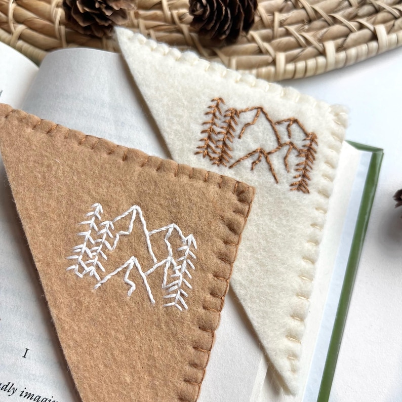 May include: Two triangular felt bookmarks. One is tan with white mountain and tree embroidery. The other is off-white with brown mountain and tree embroidery. Both are stitched around the edges and are placed in an open book.