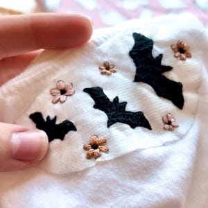 K&ouml;nnte beinhalten: Ein wei&szlig;es Kleidungsst&uuml;ck aus Stoff mit handgestickten schwarzen Flederm&auml;usen und kleinen hellrosa Blumen. Die Stickerei ist auf einen zentralen Bereich des wei&szlig;en Textils konzentriert und zeigt detaillierte Stiche von drei verschiedenen Fledermaussilhouetten.