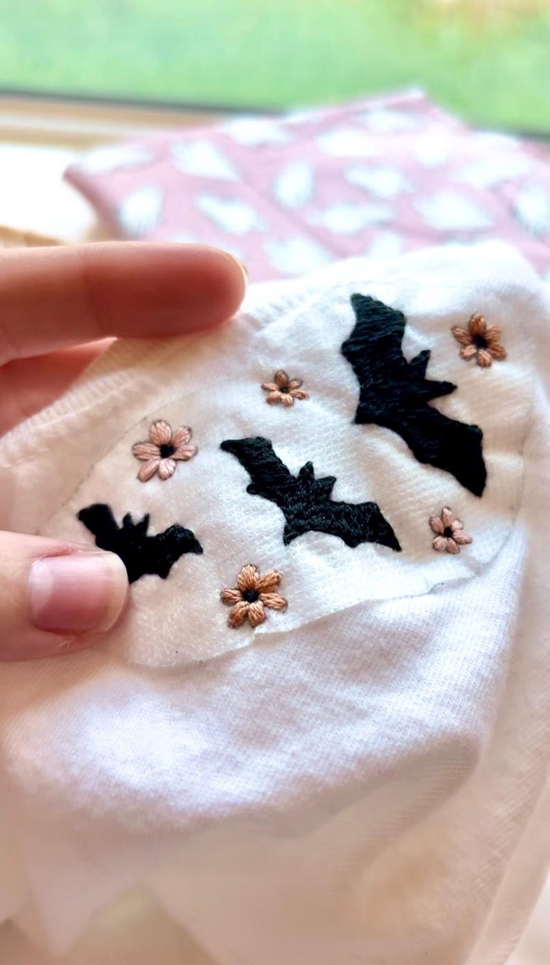K&ouml;nnte beinhalten: Ein wei&szlig;es Kleidungsst&uuml;ck aus Stoff mit handgestickten schwarzen Flederm&auml;usen und kleinen hellrosa Blumen. Die Stickerei ist auf einen zentralen Bereich des wei&szlig;en Textils konzentriert und zeigt detaillierte Stiche von drei verschiedenen Fledermaussilhouetten.