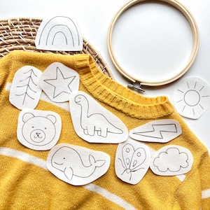 May include: A set of 10 iron-on patches featuring various designs, including a rainbow, a star, a dinosaur, a bear, a whale, a lightning bolt, a butterfly, a cloud, a sun, and a tree. The patches are white and are placed on a yellow sweater.